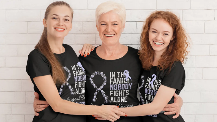 Three women wearing matching esophageal cancer awareness T-shirts with periwinkle ribbons, smiling and embracing in front of a white brick wall