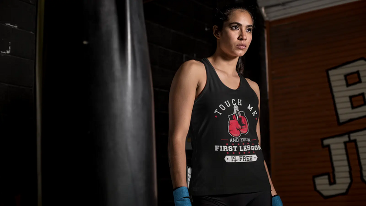 Serious female athlete wearing a black boxing-themed tank top standing near a punching bag in a dimly lit gym